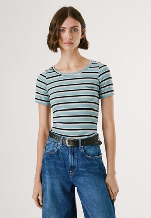 Jeune femme aux cheveux bruns courts portant un t-shirt rayé bleu, noir et blanc rentré dans un jean bleu taille haute avec une ceinture noire.