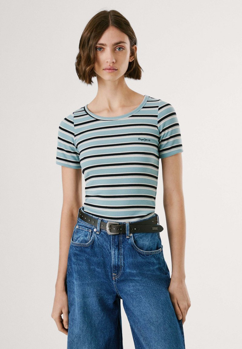 Young woman with short brown hair wearing a blue, black, and white striped t-shirt tucked into high-waisted blue jeans with a black belt.
