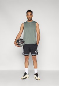 Πράσινο αμάνικο αθλητικό μπλουζάκι, μαύρο σορτς με λευκές λεπτομέρειες, κρατώντας μια μπάλα μπάσκετ. Τα παπούτσια έχουν κρεμ και μαύρο χρώμα.