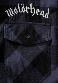 Camicia di flanella a quadri neri e blu con una piccola tasca frontale e logo ricamato "Motörhead" sopra di essa. Presenta un singolo bottone nero.