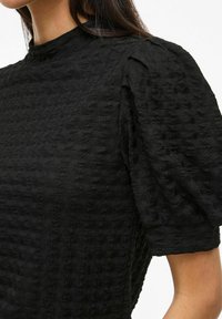 Top nero testurizzato con collo alto e maniche a sbuffo. Il tessuto presenta un sottile motivo ondulato, che ne esalta l'aspetto tridimensionale.