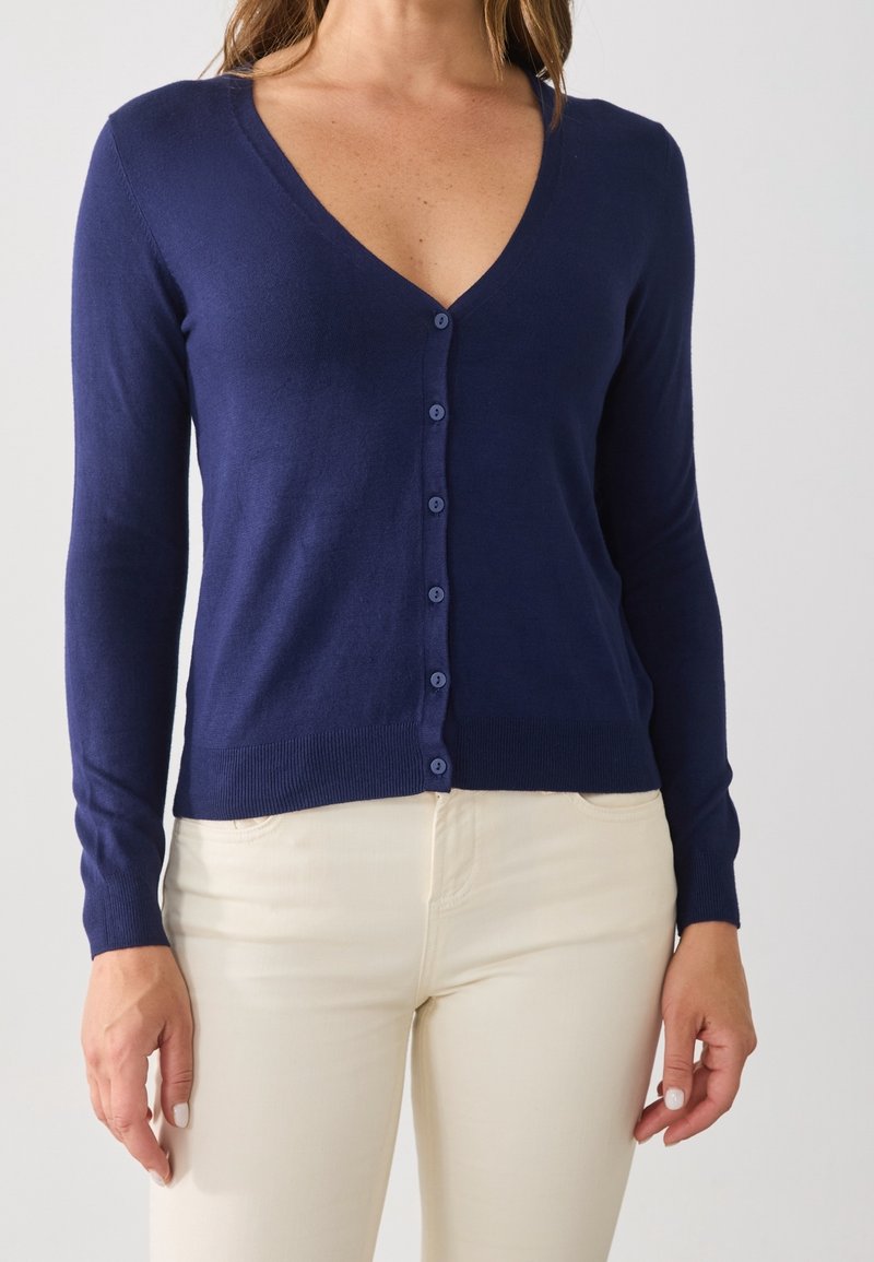 Tmavě modrý kardigan z měkké látky s hlubokým výstřihem do V, knoflíkovými detaily a dlouhými rukávy, kombinovaný se světlými kalhotami.