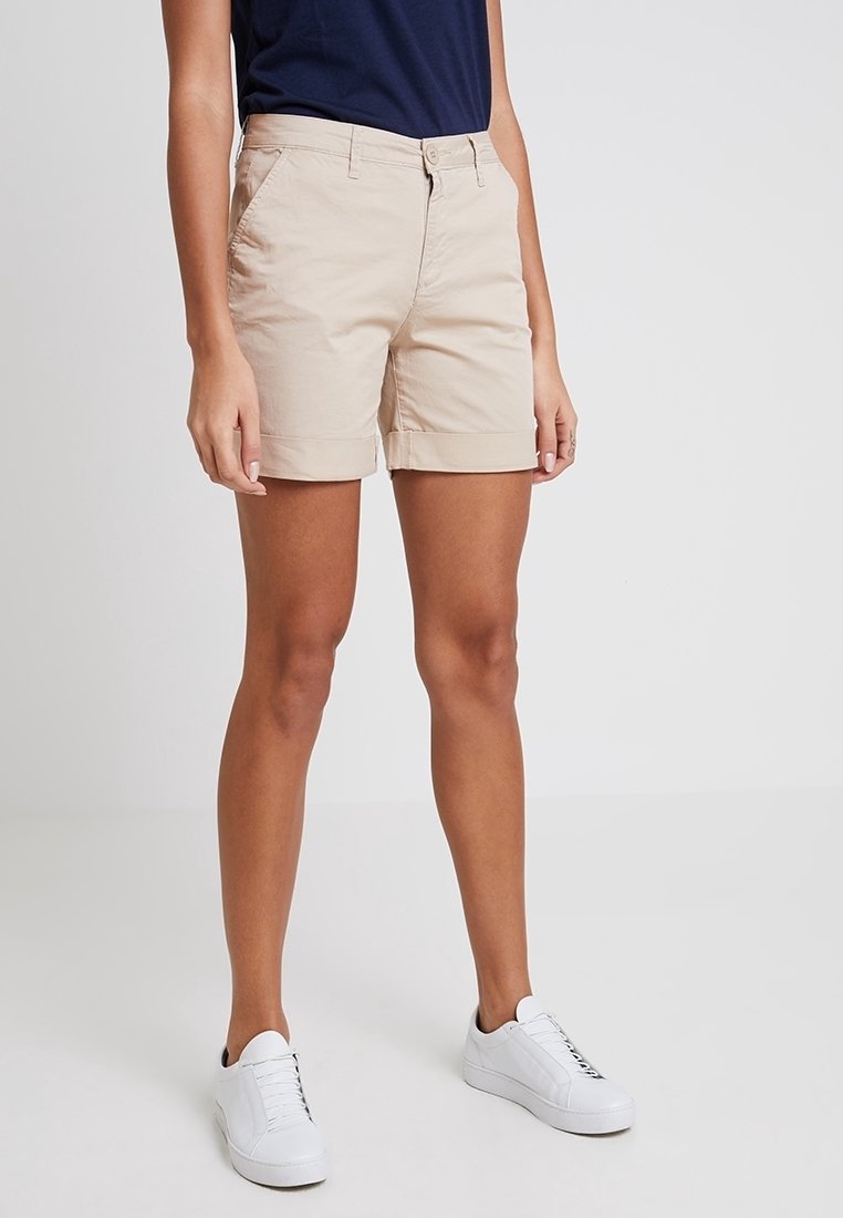 Femme portant un short beige longueur genou, un haut bleu marine et des baskets blanches, debout devant un fond clair uni.