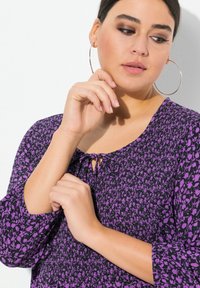 Lila Bluse mit floralen Muster, vorgemusterter Gestaltung, runder Halslinie mit Schlüssellochtiefe und weit ausgestellten Ärmeln auf einem schlichten Hintergrund.