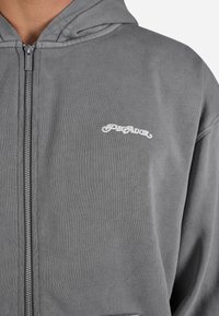 Grå zip-up hoodie lavet af en bomuldsblanding med en tekstureret finish. Har et hvidt logo på brystet og en hætte med snore.