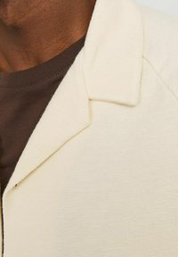 Koszula z krótkim rękawem i kołnierzykiem w jasnym kremowym kolorze, o teksturowanej powierzchni, noszona na ciemnobrązowej koszulce typu crew neck.