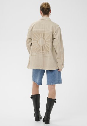 Personne avec les cheveux en chignon portant une veste beige ornée d'un motif de soleil et de texte, un short en jean bleu longueur genoux, et des bottes noires montant jusqu'au genou.