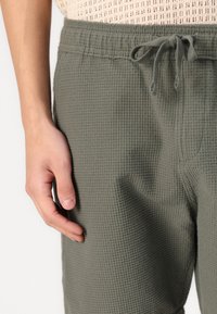 Hlače na jogger s teksturom vafla u maslinastoj zelenoj boji s elastičnom waistline i vezicama, prikazane u krupnom planu s rukom koja drži tkaninu.