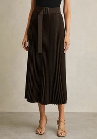 Gonna midi plissettata marrone con una fascia in vita ampia e una cintura, texture liscia e silhouette fluida. Indossata con tacchi nudi con cinturini.