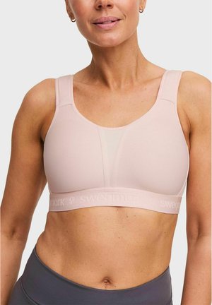 Vrouw met een lichtroze sportbeha met brede bandjes en grijze, hooggetailleerde legging, getoond vanaf halverwege de borst tot de taille.