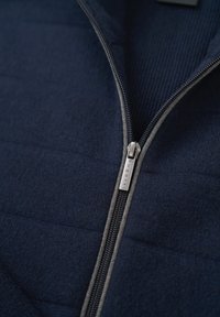 Navyblaue Zip-Jacke aus strukturiertem Stoff, mit einem silbernen Reißverschluss und gerippten Akzenten. Gestepptes Design in Abschnitten sichtbar.
