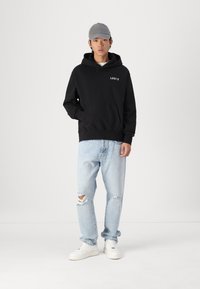 Felpa nera con logo bianco "LEVI'S", indossata con jeans strappati blu chiaro e scarpe da ginnastica bianche. Il modello indossa anche un cappellino grigio. Design minimalista.