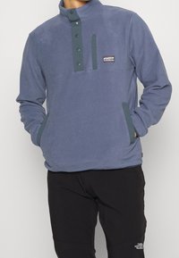 Fleece-pullover i mörkblått med hög krage, grön knappplacket och en bröstficka; har två sidofickor. Mjuk textur.