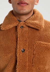 Veste en polaire marron avec col, dotée d'une fermeture à boutons et d'une poche à rabat. Tissu texturé avec une apparence douce et confortable.