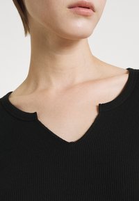 Schwarzes, geripptes Oberteil mit einem V-Ausschnitt und einer rohen Kante. Der Stoff ist strukturiert und betont das taillierte Design um den Kragen.