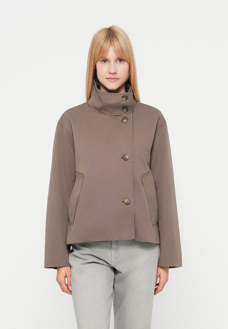 Femme debout face à l'avant portant une veste taupe à col montant boutonnée et un pantalon gris clair sur un fond blanc uni.