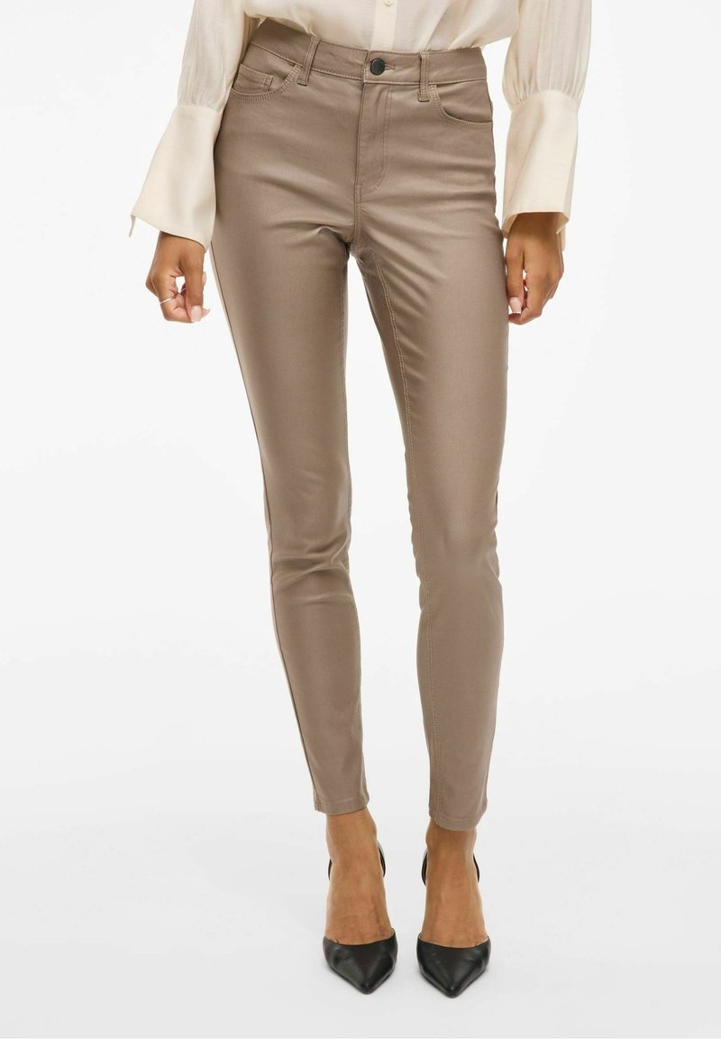 Femme portant un pantalon slim taupe, un chemisier crème à manches longues et des talons pointus noirs, debout devant un fond blanc.