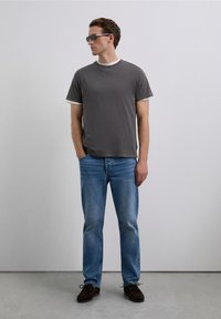 Homme portant un t-shirt superposé gris foncé, un jean bleu clair, des chaussures noires et des lunettes de soleil, se tenant contre un mur clair et uni.