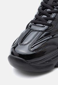 Fekete lakkbőr sportcipő ívelt, lekerekített orral, texturált fűzőkkel és kontrasztos gumitalppal. Karcsú kialakítás fényes felülettel.