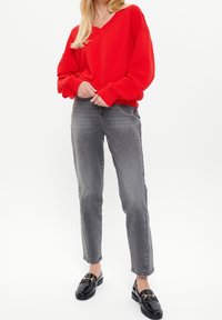 Vrouw staat rechtop en draagt een rode V-hals trui, grijze versleten jeans en zwarte loafers met gouden accenten op een witte achtergrond.