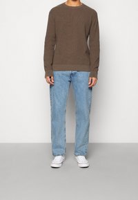 Brun texturerad långärmad tröja kombinerad med ljusblå raka jeans och vita sneakers, stående mot en enfärgad bakgrund.