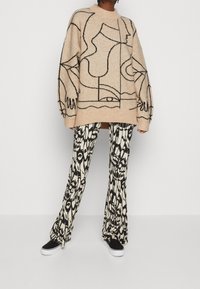 Beige Strickpullover mit schwarzem abstraktem Liniendesign, kombiniert mit schwarz-weißen gemusterten Schlaghosen und schwarzen Sneakers.