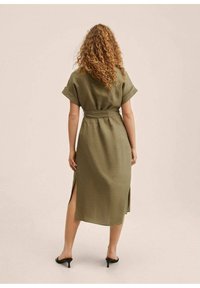 Olivengrøn linnedkjole med smal talje, korte opsmøgede ærmer og sideslids, der når til midt på læggen, fremhævet af højhælede sko.