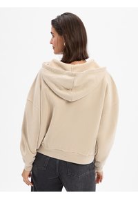 Overmaat beige hoodie van zacht, gestructureerd stof, met een grote capuchon en geribde manchetten. Draagt met donkergrijze jeans, wat een relaxte pasvorm laat zien.