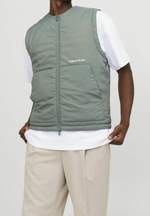 Mann trägt salbegrüne ärmellose Weste mit Reißverschluss über weißem T-Shirt und beiger Hose, Hände halten den Saum des T-Shirts.