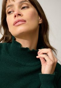 Maglione verde a collo alto realizzato in morbido materiale a maglia, con colletto a onde e polsini a costine, caratterizzato da una texture liscia.