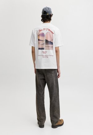 Osoba ubrana w biały T-shirt z grafiką przedstawiającą zachód słońca i falę na plecach, szare dżinsy, brązowe buty i szarą czapkę z daszkiem, stojąca tyłem.