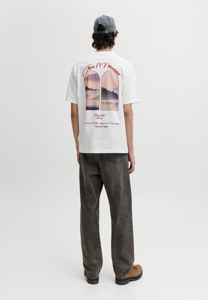 Personne portant un t-shirt blanc graphique avec un dessin de coucher de soleil et de vague dans le dos, un jean gris, des bottes marron clair et une casquette de baseball grise, debout de dos.