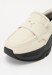 Bottines Puma à enfiler avec un dessus en cuir crème, semelle en caoutchouc noire, sangle de maintien, et un petit logo accentué sur le côté.