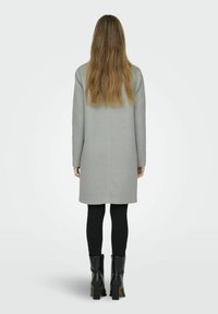Lichtgrijze lange mouwenjas met een rechte snit, voorzien van een naad aan de achterkant. Zwarte leggings en zwarte kniehooglaarzen maken de look compleet.