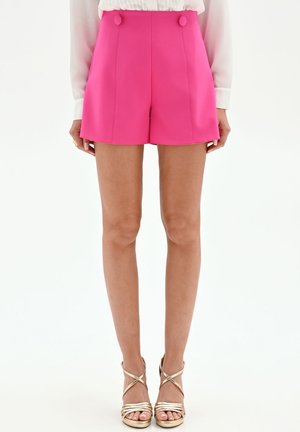 Femme portant un short taille haute rose vif, cintré avec des boutons recouverts de tissu, une blouse blanche et des sandales à talons dorées métalliques à brides.