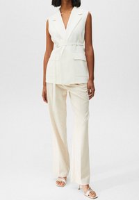 Biały bezrękawnik z wiązaniem w talii i dwiema kieszeniami z przodu, zestawiony z kremowymi szerokimi spodniami i sandałami na szpilce. Gładka tekstura materiału.
