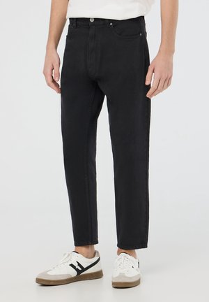 Mand iført sorte jeans med lige ben og hvide sneakers med sorte detaljer, stående foran en ensfarvet baggrund.