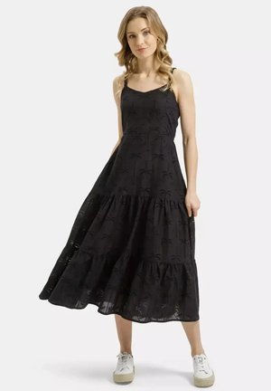 Kobieta ubrana w czarną, bezrękawnikową sukienkę sięgającą do kostek z detalami ażurowymi, stojąca i trzymająca spódnicę, w połączeniu z białymi trampkami.