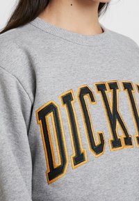 Sweat-shirt gris avec col côtelé et "DICKIES" brodé en grandes lettres noires et jaunes. Tissu doux et texturé.
