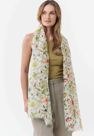 Frau trägt einen langen, floralen Schal mit Pastellblumen über einem olivgrünen Oberteil und beigefarbenen Hosen, steht vor einem weißen Hintergrund.