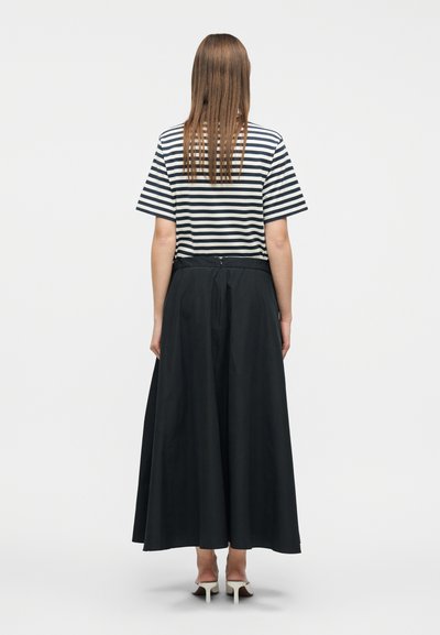 Γυναίκα με μακριά ίσια μαλλιά που φοράει μαύρο και λευκό ριγέ κοντομάνικο πουκάμισο, μακριά μαύρη φούστα και λευκά παπούτσια με τακούνι, και στέκεται με την πλάτη στραμμένη.