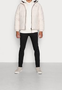 Gewatteerde jas in lichtroze met een capuchon, voorzien van een rits aan de voorkant, elastische manchetten en een witte hoodie daaronder. Gekleed met zwarte jeans en lichtgekleurde schoenen.