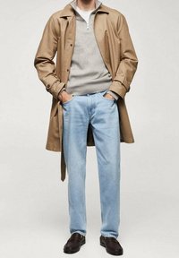 Hnědý trenčkot přes šedý hřejivý svetr s ribovaným vzorem, světle modré džíny a tmavě hnědé boty. Oblečení má zipový límec a volný střih.