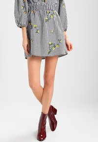 Schwarz-weißes Gingham-Kleid mit gelber Blumenstickerei, elastischem Bund und Ballonärmeln. Getragen mit bordeauxfarbenen Knöchelschuhen.