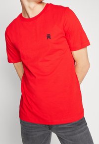 T-shirt en coton rouge à manches courtes, col rond, avec un petit logo bleu marine sur la poitrine. Associé à un jean gris foncé.