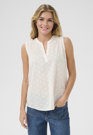 Blouse blanche sans manches avec un décolleté en V, texturée de petits motifs en forme de cœur, en tissu léger et transparent. Associée à un jean en denim bleu.