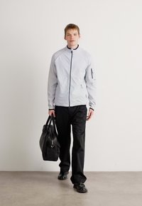 Jachetă zip-up de culoare gri deschis cu accente negre, pantaloni negri și o geantă neagră. Textură netedă, design modern, croială slim.