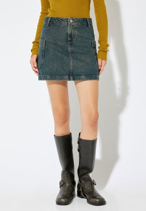 Jupe mini en denim bleu foncé avec deux poches latérales, associée à un haut ajusté à manches longues jaune moutarde et des bottes noires en cuir hautes.