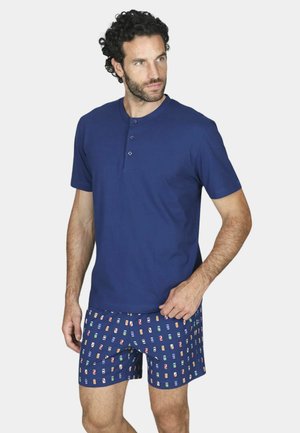 Uomo con capelli ricci che indossa una camicia blu a maniche corte abbottonata e pantaloncini blu navy con motivi colorati di lattine, che guarda di lato.
