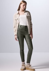 Olivgrönt åtsittande byxor med en glansig yta, kombinerat med ett vitt linne och en beige skjorta med knappar. Mysiga beige skor och grå strumpor.